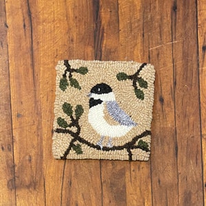 May include: A beige and brown rug with a tufted design of a bird perched on a branch with green leaves. The bird is white with a black head and gray wings.