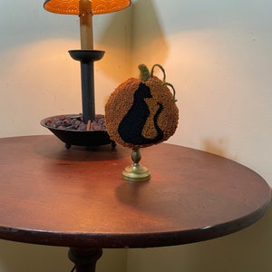 Op de afbeelding: Een bruine, pompoenvormige naaldkunst met een zwart katten silhouet op een gouden standaard. De pompoen staat op een houten tafel naast een lamp met een bruine kap.