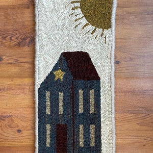 May include: A hand-stitched rug depicting a blue house with a brown roof and a yellow star on the front. The house is set against a white background with a yellow sun in the top right corner.