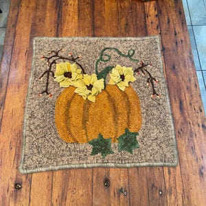 May include: A square, handmade rug featuring a large orange pumpkin with yellow flower accents and green leaves. The rug has a brown background and a brown border, perfect for fall decor.