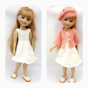 May include: Two dolls wearing white dresses with ruffled hems and white flower embellishments. One doll is wearing a pink knitted cardigan and a pink beret.