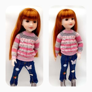 Può includere: Una bambola con lunghi capelli rossi che indossa un maglione a righe rosa e grigio e jeans blu con decorazioni a farfalla e fiori.