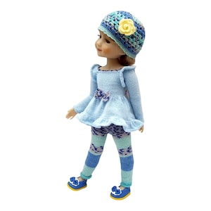 Puede incluir: Una muñeca vestida con un conjunto de punto azul claro, que incluye un vestido con lazo, leggings y un sombrero con una flor amarilla. La muñeca también lleva zapatos azules con lazos. El conjunto está hecho a mano.