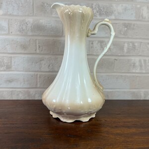 May include: A vintage ceramic pitcher with a cream and tan color gradient. The pitcher has a slender neck, a bulbous base, and a curved handle. The rim is scalloped, and the base has a decorative floral design.