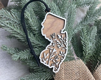May include: A wooden ornament shaped like the state of New Jersey with a white floral design cut out. The ornament has a black string for hanging.