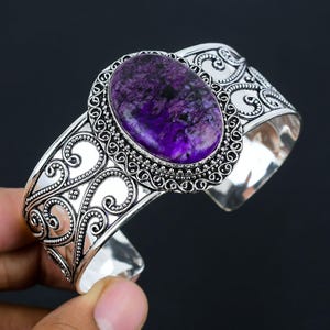 Pode incluir: Uma pulseira de punho prateada com uma pedra preciosa ovalada, roxa escura. A pedra preciosa está engastada numa moldura decorativa em estilo filigrana. A pulseira tem um design aberto e é segurada por uma mão contra um fundo escuro.