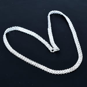 Op de afbeelding: Een zilverkleurige ketting met een geweven ontwerp. De ketting wordt getoond tegen een zwarte achtergrond, waarbij de ingewikkelde schakels en sluiting worden benadrukt. De ketting is gemaakt van een glanzend, reflecterend metaal.