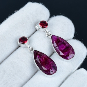 May include: A pair of silver dangle earrings featuring teardrop-shaped ruby gemstones. Each earring has a smaller round ruby gemstone at the top of the teardrop.
