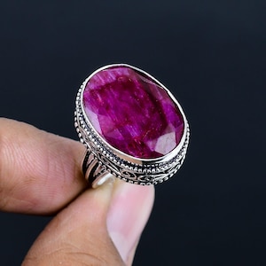 May include: A silver ring featuring a large, oval-cut ruby gemstone. The ruby is a deep, vibrant pink-red color. The ring has an ornate, detailed setting with a textured band.