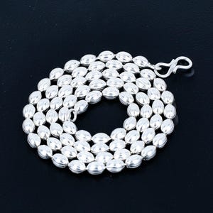 Puede incluir: Un collar plateado con una serie de cuentas ovaladas. Las cuentas están ensartadas para formar una cadena continua. El collar tiene un cierre. El fondo es negro. Longitud aproximada: 45 cm.