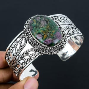May include: A silver cuff bracelet with an oval-shaped green and pink gemstone set in a decorative silver bezel. The bracelet has a filigree design.