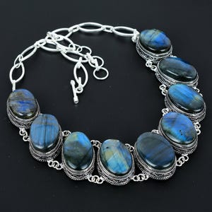 May include: A silver necklace featuring oval-shaped labradorite stones with blue and green iridescence. The stones are set in ornate silver bezels and connected by a silver chain with a toggle clasp. The necklace is displayed on a black surface.