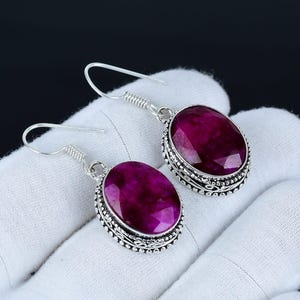 Può includere: Orecchini in argento con grandi pietre preziose ovali color magenta. Le pietre sfaccettate sono incastonate in montature d'argento dettagliate. Gli orecchini sono mostrati su uno sfondo bianco, con uno sfondo scuro.