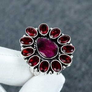 May include: A silver ring featuring a large, teardrop-shaped, deep red gemstone at the center, surrounded by eleven oval-shaped, smaller red gemstones. The ring is set against a neutral background.
