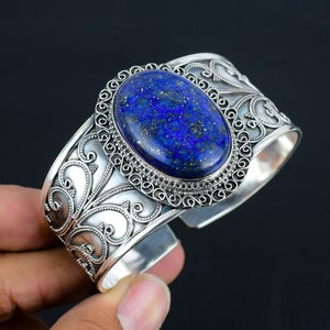 Pode incluir: Um bracelete de punho de prata com um design ornamentado apresenta uma grande pedra oval de lapis-lazúli. A pedra é de cor azul escura com manchas douradas.