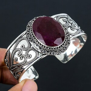 May include: A silver cuff bracelet with an ornate design featuring a large oval-shaped ruby gemstone.