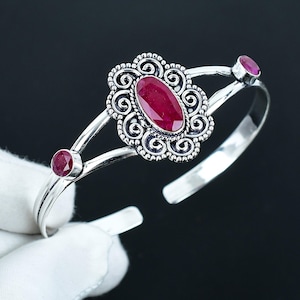 May include: A silver-toned cuff bracelet featuring a large oval ruby gemstone at the center, surrounded by an ornate, swirling design. Two smaller ruby stones are set on either side of the bracelet. The bracelet is open-ended.