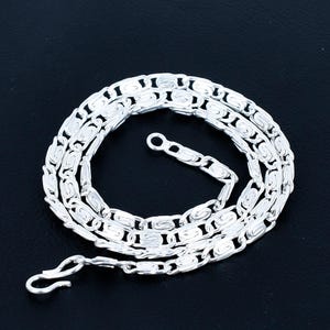 Puede incluir: Un collar de cadena plateada con un diseño de eslabones texturizados único. El collar se encuentra sobre una superficie negra, mostrando su intrincado patrón y el cierre de gancho. Los eslabones de la cadena tienen una forma ovalada alargada con una superficie texturizada detallada.