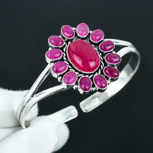 May include: A silver bangle bracelet with a pink gemstone center surrounded by smaller pink gemstones. The bracelet is open and has a simple design.