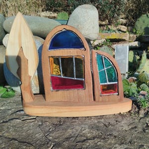Könnte beinhalten: Eine Holzskulptur mit einem stilisierten Haus mit Buntglasfenstern in Blau, Rot, Gelb und Grün. Neben dem Haus befindet sich eine hölzerne Baumform. Die Skulptur steht auf einer Steinoberfläche.