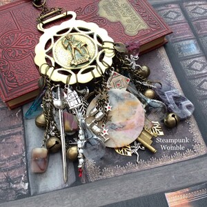 May include: A brass charm necklace with a variety of charms, including a sword, a key, a bell, and a stone. The necklace is hanging from a book with the text "Stories of the Day & Night".