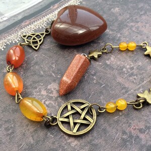 May include: A brown heart-shaped stone, a brown and gold pendulum, and a chain with gold stars and orange and yellow beads. The chain has a gold pentacle charm and a gold Celtic knot charm.