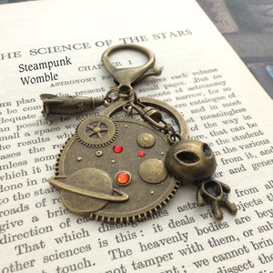 May include: A steampunk-style key chain with a bronze-colored gear design, a small rocket, a planet with rings, and an alien figure. The key chain has red gemstones and a silver key ring.