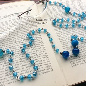May include: A silver chain necklace with blue glass beads and larger blue agate beads. The necklace is lying on a book with text about occultism.
