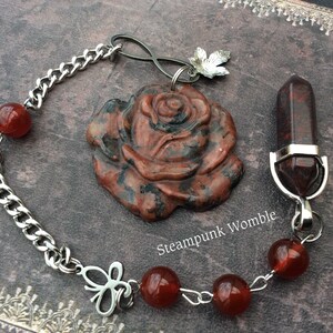 May include: A silver chain necklace with a red rose pendant, a red crystal point pendant, and red carnelian beads. The rose pendant is carved from red jasper. The chain has an infinity symbol charm and a butterfly charm. The text "Steampunk Womble" is visible on the image.