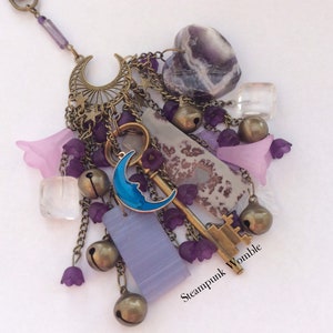 Pu&ograve; includere: Una collana con ciondoli viola e argento con una chiave, una luna, campane e cristalli. La collana ha un'etichetta "Steampunk Womble".