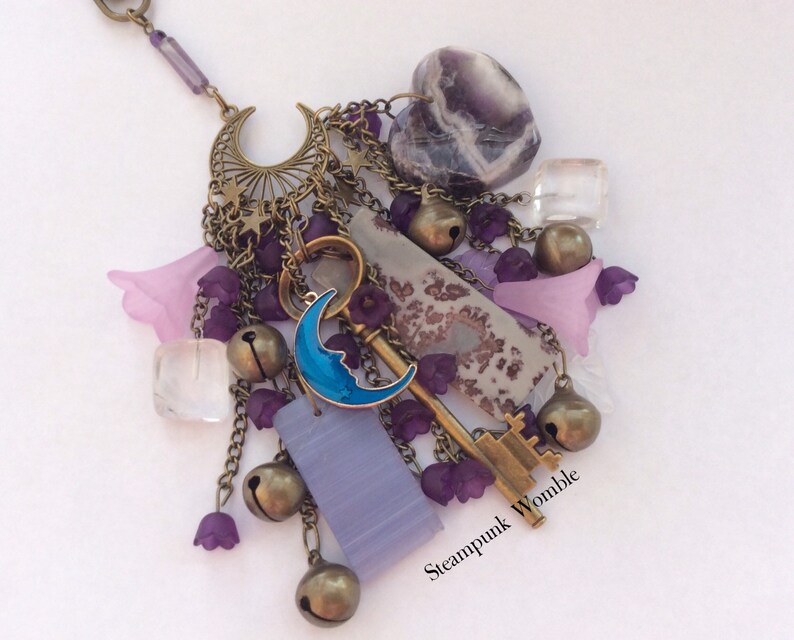 Pu&ograve; includere: Una collana con ciondoli viola e argento con una chiave, una luna, campane e cristalli. La collana ha un'etichetta "Steampunk Womble".