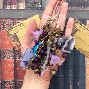 Pu&ograve; includere: Un ciondolo in stile steampunk con una luna crescente, chiavi, campane e cristalli di ametista. Il ciondolo &egrave; appeso a una catena ed &egrave; circondato da fiori viola e rosa.