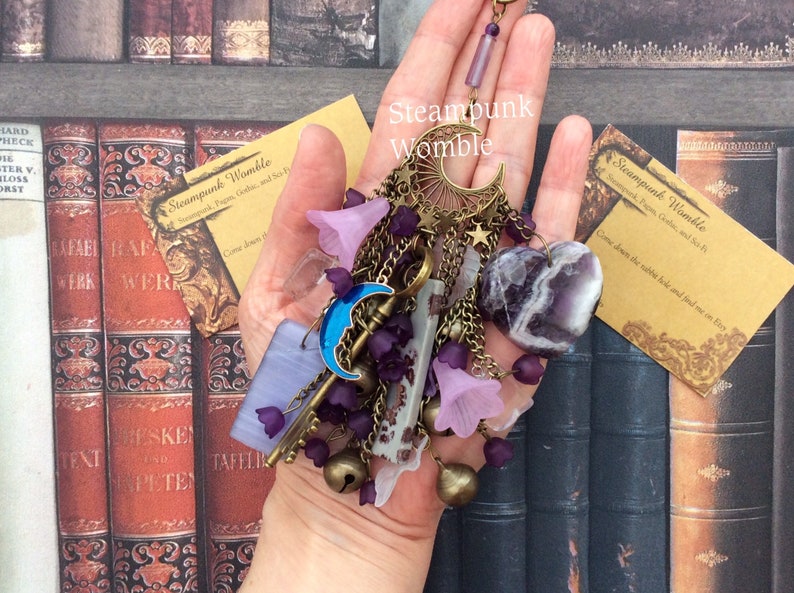Pu&ograve; includere: Un ciondolo in stile steampunk con una luna crescente, chiavi, campane e cristalli di ametista. Il ciondolo &egrave; appeso a una catena ed &egrave; circondato da fiori viola e rosa.