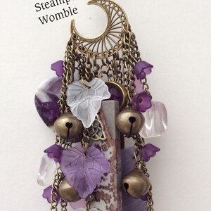 Pu&ograve; includere: Un ciondolo viola e bianco con un design a forma di luna crescente, campane, foglie e cristalli. Il ciondolo &egrave; etichettato "Steampunk Womble" e "Back View".