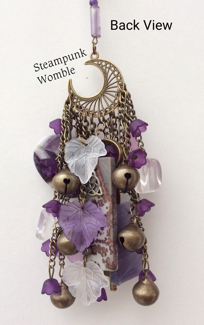 Pu&ograve; includere: Un ciondolo viola e bianco con un design a forma di luna crescente, campane, foglie e cristalli. Il ciondolo &egrave; etichettato "Steampunk Womble" e "Back View".
