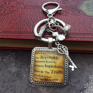 Può includere: Un portachiavi color argento con un ciondolo quadrato recante il testo: When You Have Eliminated The Impossible, Whatever Remains, However Improbable, Must Be The Truth. Sherlock Holmes, The Sign Of Four (1890). Ciondoli: pipa, violino, chiave, torre.