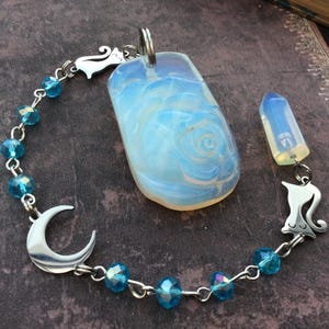 May include: A celestial-themed pendant and bracelet set. The pendant features a large, blue-swirled stone, and a smaller crystal. The bracelet has blue beads, a crescent moon charm, and cat-shaped connectors. The set is displayed on a textured, dark surface.