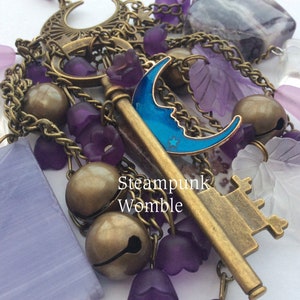 Pu&ograve; includere: Un ciondolo a tema steampunk con una chiave, campane e varie perline di vetro viola e trasparente. La chiave ha un ciondolo a forma di mezzaluna blu attaccato. Il testo "Steampunk Womble" &egrave; visibile nell'immagine.