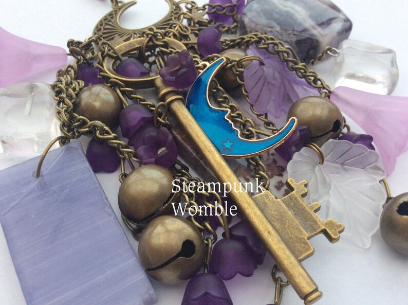 Pu&ograve; includere: Un ciondolo a tema steampunk con una chiave, campane e varie perline di vetro viola e trasparente. La chiave ha un ciondolo a forma di mezzaluna blu attaccato. Il testo "Steampunk Womble" &egrave; visibile nell'immagine.