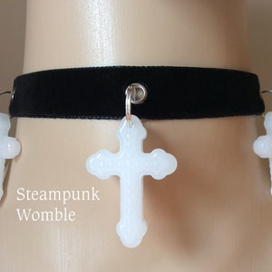 Può includere: Choker in velluto nero con tre ciondoli a forma di croce bianca. Il choker ha una chiusura in metallo argentato e le croci sono attaccate con anelli argentati.
