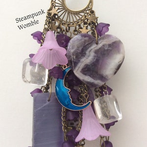 Pu&ograve; includere: Un ciondolo a forma di cuore in ametista viola con un ciondolo a forma di luna crescente blu, cristalli di quarzo trasparenti e ciondoli a forma di fiore viola appesi a una catena in bronzo con una luna crescente e stelle.