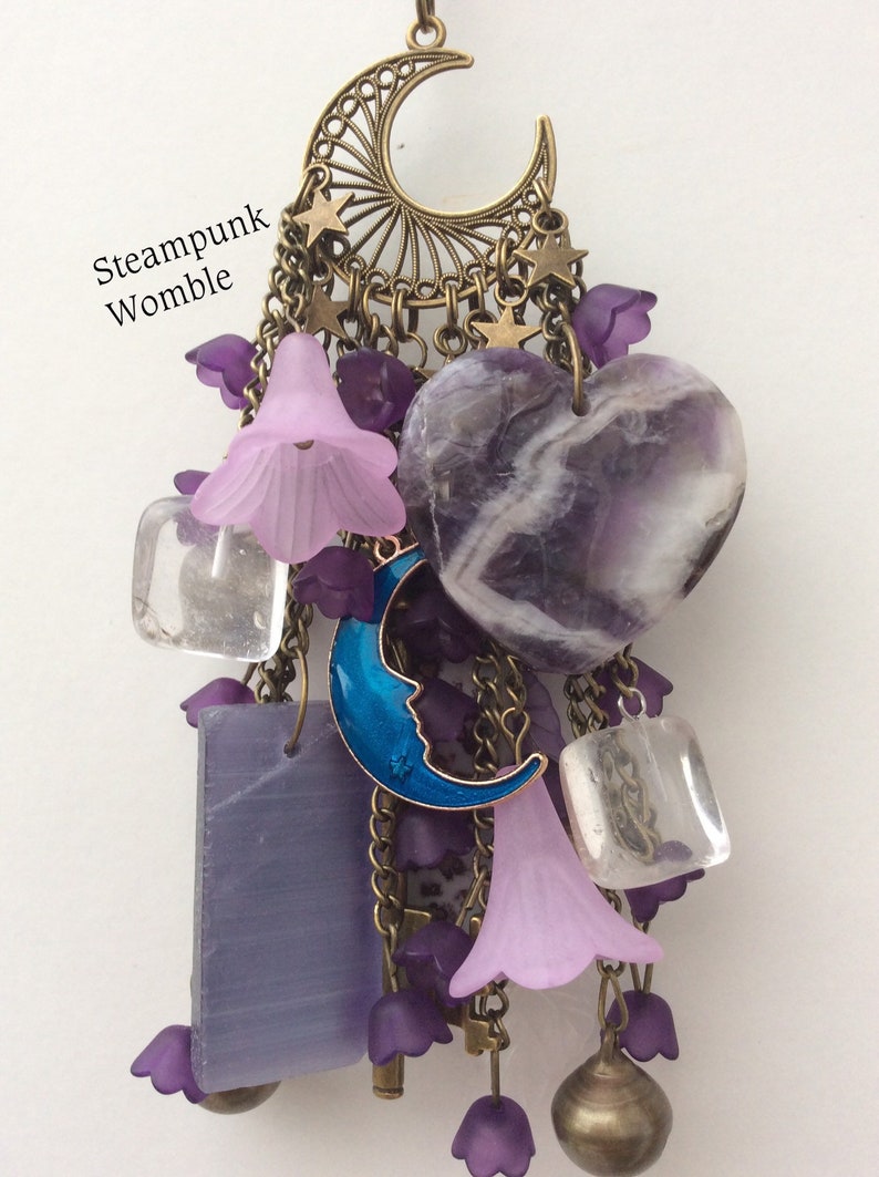 Pu&ograve; includere: Un ciondolo a forma di cuore in ametista viola con un ciondolo a forma di luna crescente blu, cristalli di quarzo trasparenti e ciondoli a forma di fiore viola appesi a una catena in bronzo con una luna crescente e stelle.