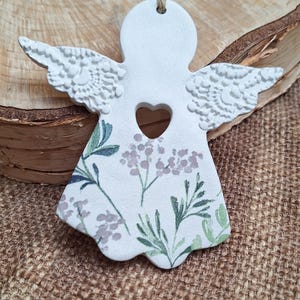 May include: White ceramic angel ornament with a heart-shaped cutout and floral design. The angel has textured wings and a jute string for hanging. The floral pattern includes green and lavender accents.