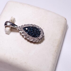 May include: A teardrop-shaped pendant crafted from polished silver-toned metal, featuring a central cluster of dark blue stones surrounded by a halo of small, clear, sparkling stones. The pendant includes a sturdy bail for attachment to a chain.