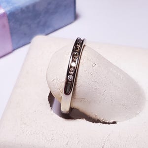 May include: A silver ring with multiple small, clear gemstones set in a row. The ring is displayed on a white, textured surface. A small, blue gift box with a lavender ribbon is in the background.