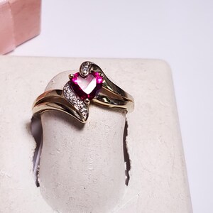 May include: A gold ring with a heart-shaped, ruby-colored stone and small white accent stones. The ring is displayed on a white surface, with a pink gift box in the background. A romantic jewelry piece.