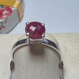 May include: A silver ring with a red gemstone. The ring is on a white display stand.