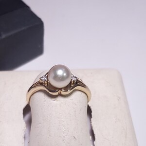 May include: A gold ring featuring a central white pearl flanked by two small clear stones. The band has a smooth, polished finish and a curved setting that holds the pearl securely in place, creating a classic look for this piece of jewelry.