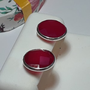 May include: A pair of oval-shaped stud earrings. Each earring features a faceted, deep red gemstone set in a silver-colored metal bezel. The earrings are displayed on a white surface, with a floral patterned box in the background.