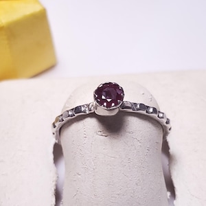 May include: A silver ring with a round, dark red gemstone set in a bezel. The band features a textured design. A yellow gift box is partially visible in the background. The ring is displayed on a white surface.